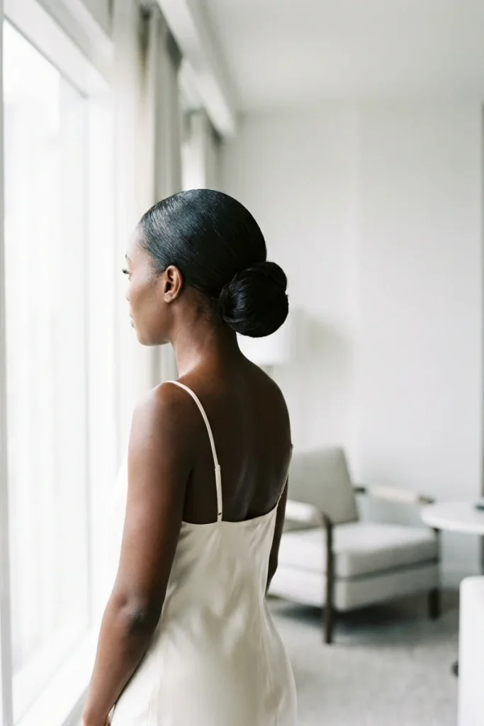 Bride with a sleek low bun at the nape and an ivory satin slip dress, looking out of a bright hotel window