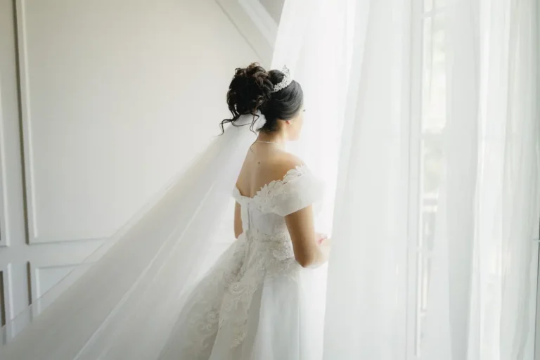 Elegant bridal updo with pearl tiara and cathedral veil, back view against soft white curtain background