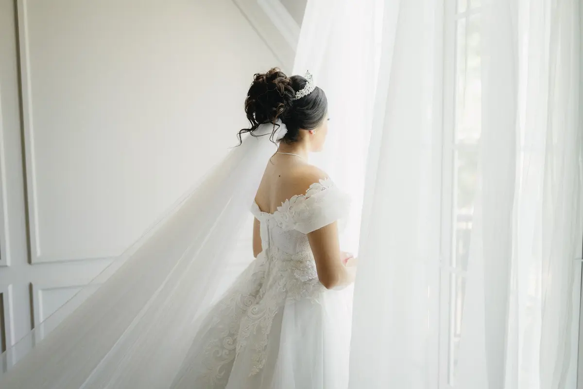 Elegant bridal updo with pearl tiara and cathedral veil, back view against soft white curtain background