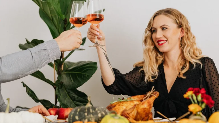 Woman with soft blonde waves raising a glass at a Thanksgiving table with roast turkey and pumpkins
