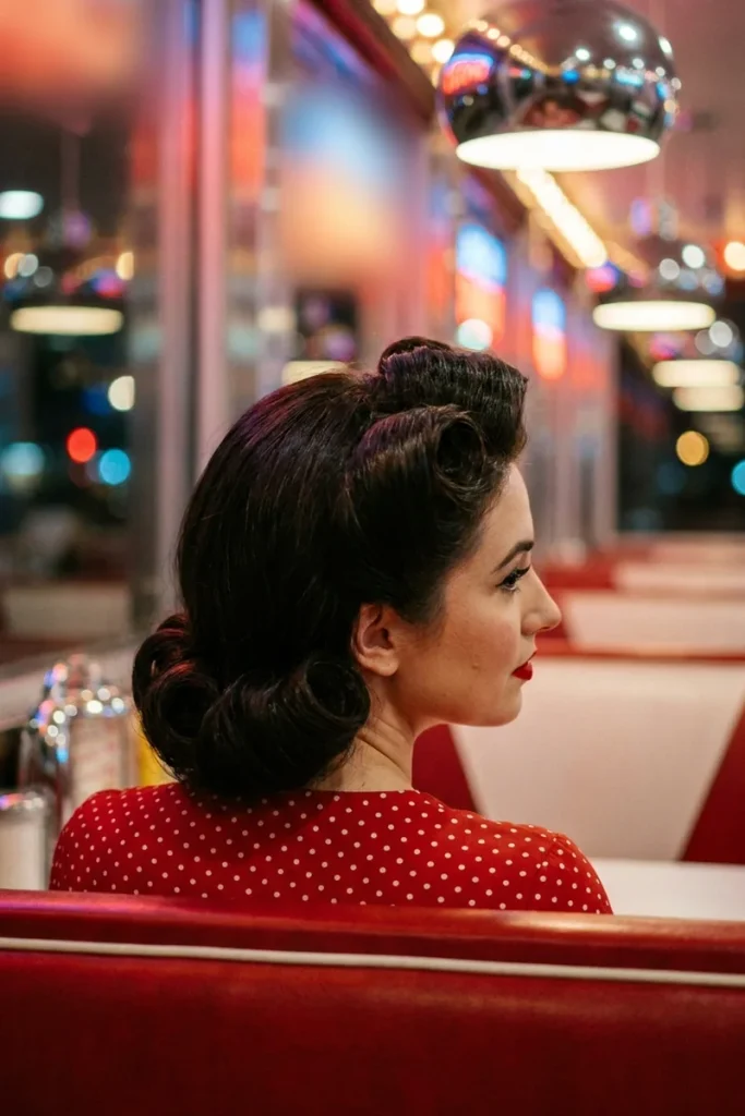 Victory-roll updo on long jet black hair with glossy rolls on top and a low roll at the nape, woman in a red polka-dot dress at a retro diner