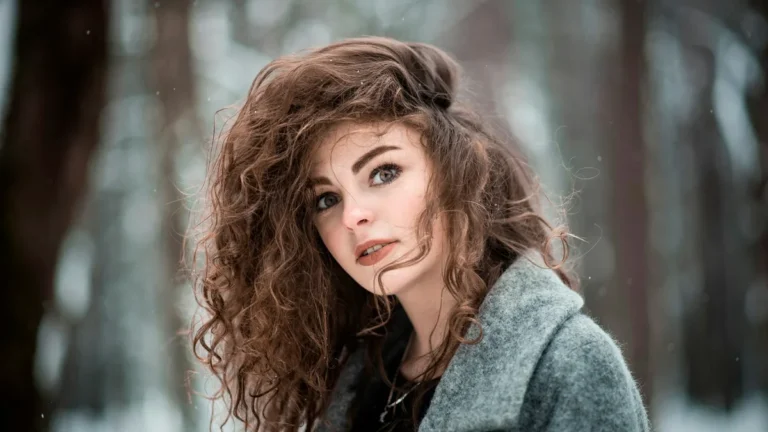 Woman with voluminous curly chestnut brown hair in a gray wool coat in a snowy forest