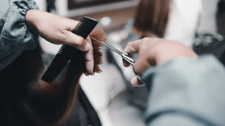 Hairstylist in a salon cape holding the ends of long auburn hair between comb and fingers and trimming the tips with scissors