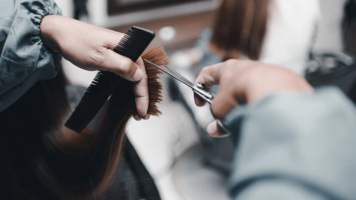 Hairstylist in a salon cape holding the ends of long auburn hair between comb and fingers and trimming the tips with scissors