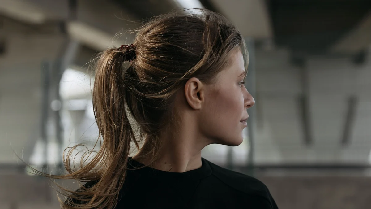 Side profile of a woman with a mid-height textured ponytail tied with a small hair tie, loose strands moving in the breeze