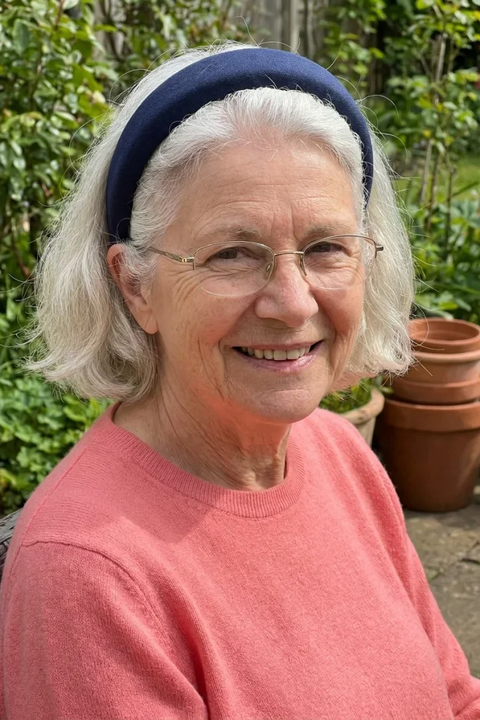 Smiling woman in her 60s with short silver hair pushed back by a wide navy padded headband, wearing glasses in a garden